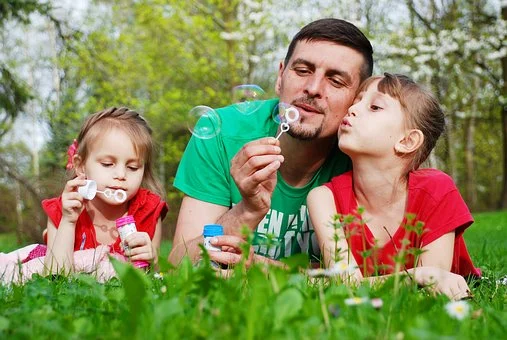 image of father and two daughters resting on grass and blowing bubbles together